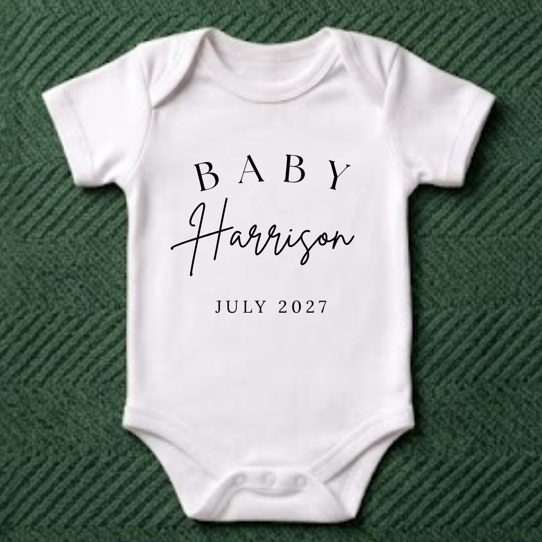A white short-sleeve baby bodysuit laid flat on a dark green textured fabric background. The bodysuit has black printed text reading 'BABY Harrison JULY 2027' on the front.