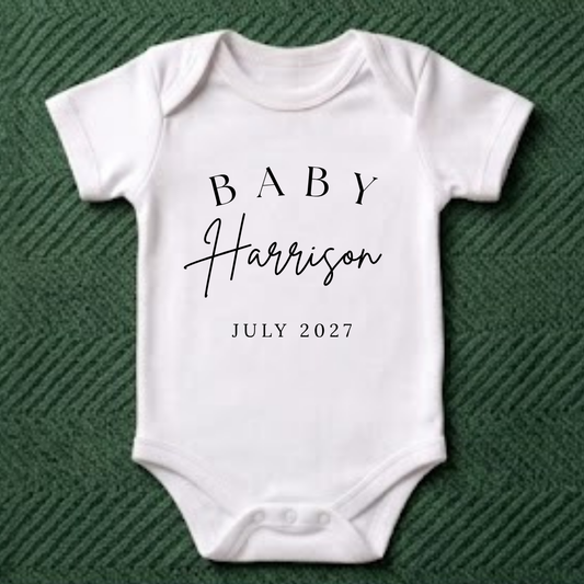A white short-sleeve baby bodysuit laid flat on a dark green textured fabric background. The bodysuit has black printed text reading 'BABY Harrison JULY 2027' on the front.